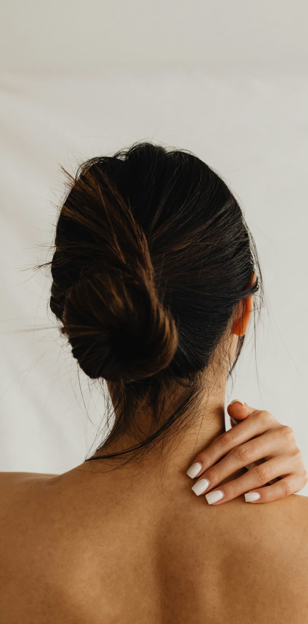 A person with dark hair tied in a bun viewed from behind, gently touching their shoulder with manicured white nails.